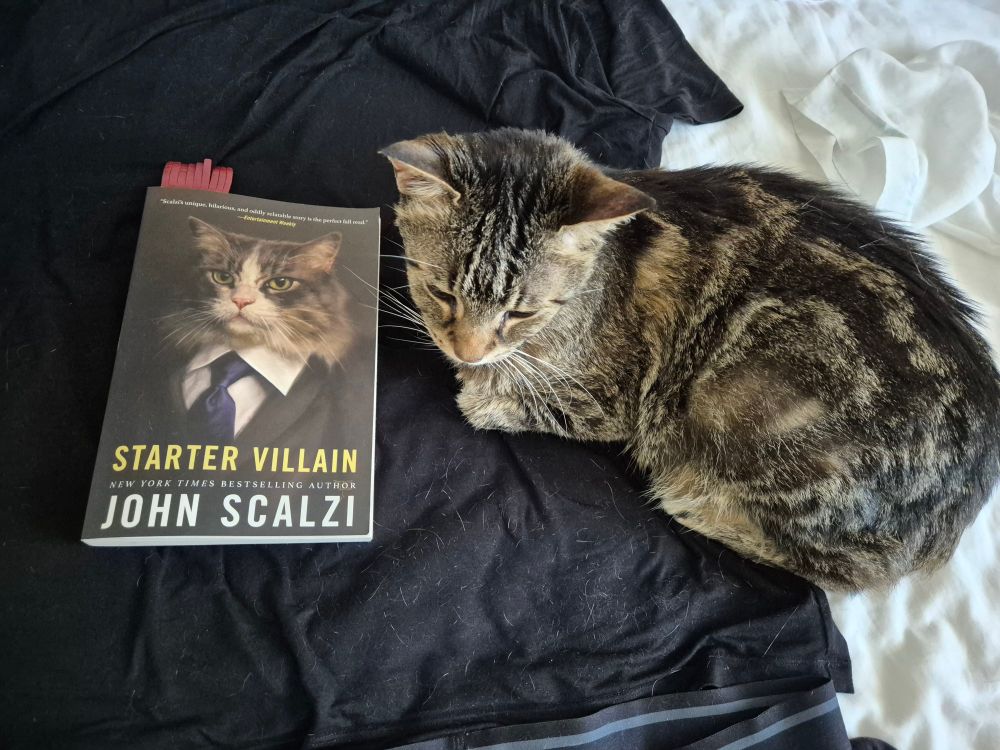 A tabby cat is lounging on her side on a dark t-shirt next to a book. The book is "Starter Villain" by John Scalzi and there is a picture of a cat in a suit on the cover. 