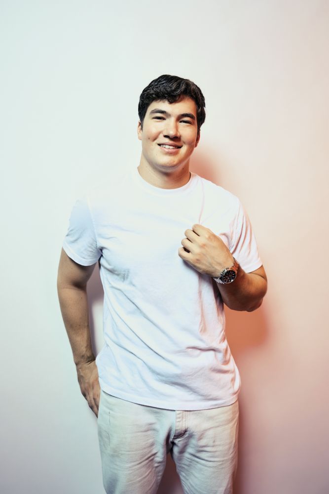 Niels Levy-Thiebaut. A student with short dark hair is wearing a white t-shirt and light khaki pants. He is wearing a silver analog watch and standing in front of a white background. Photo Credit: Matt Wright-Steel
