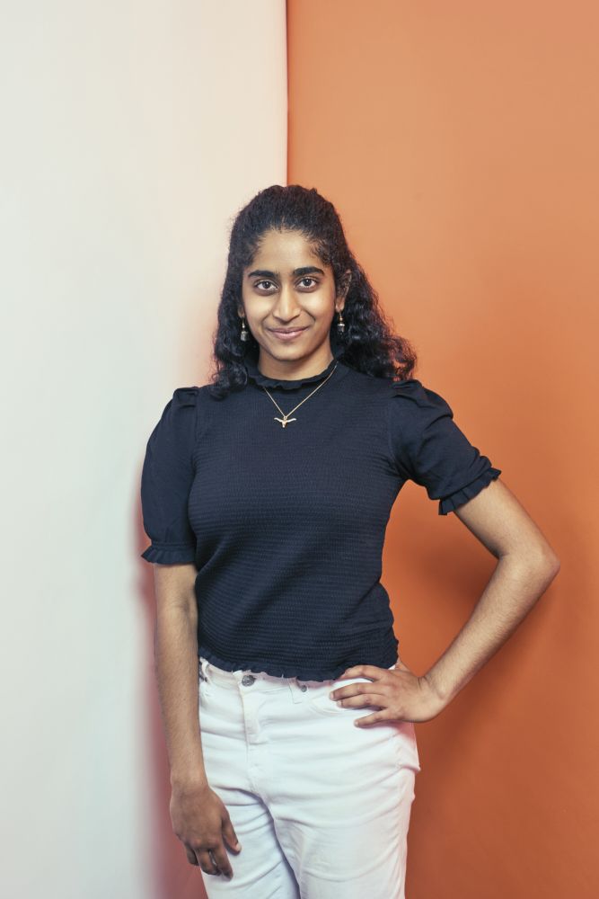 Neha Donthineni. A student with long, dark, curly hair wears a black top with white pants. She is wearing a Longhorn pendant on a gold chain and standing in front of a backdrop that is half white (left) and half burnt orange (right). Photo Credit: Matt Wright-Steel
