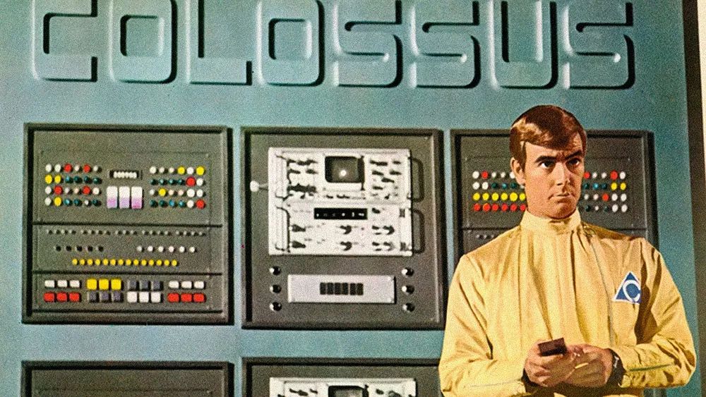 A scientist in a yellow jumpsuit stands in front of a huge panel of buttons and electronic displays of a 1970s intelligent computer.  Above the panel is the name of the computer: “Colossus”.  An image from the 1970 movie “Colossus: The Forbin Project”. 
