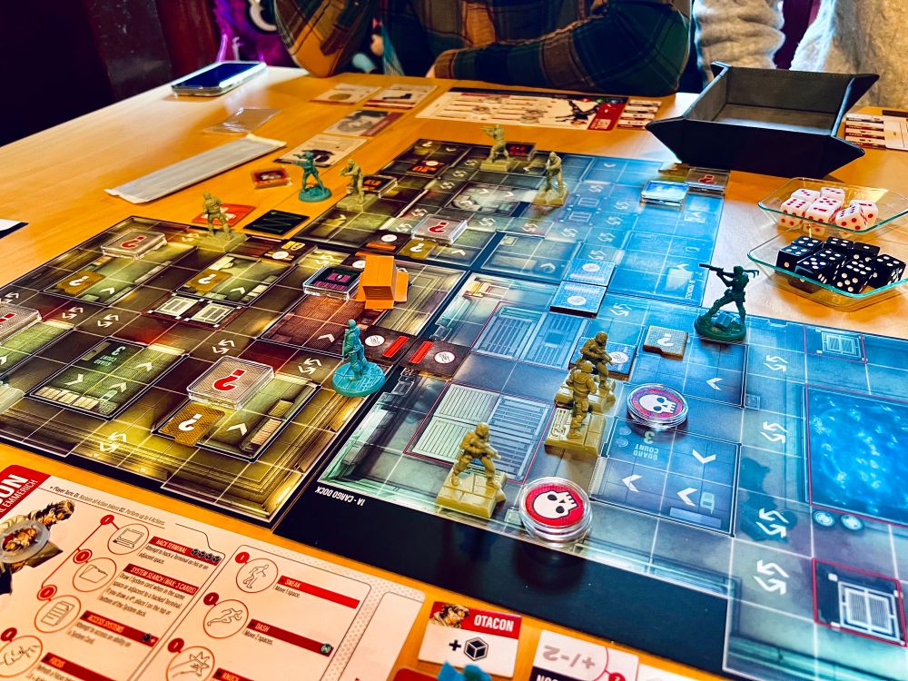 A full table set up with Metal Gear Solid the boardgame. It shows several board pieces and miniatures of the characters and guards. There is also a cardboard box miniature. 