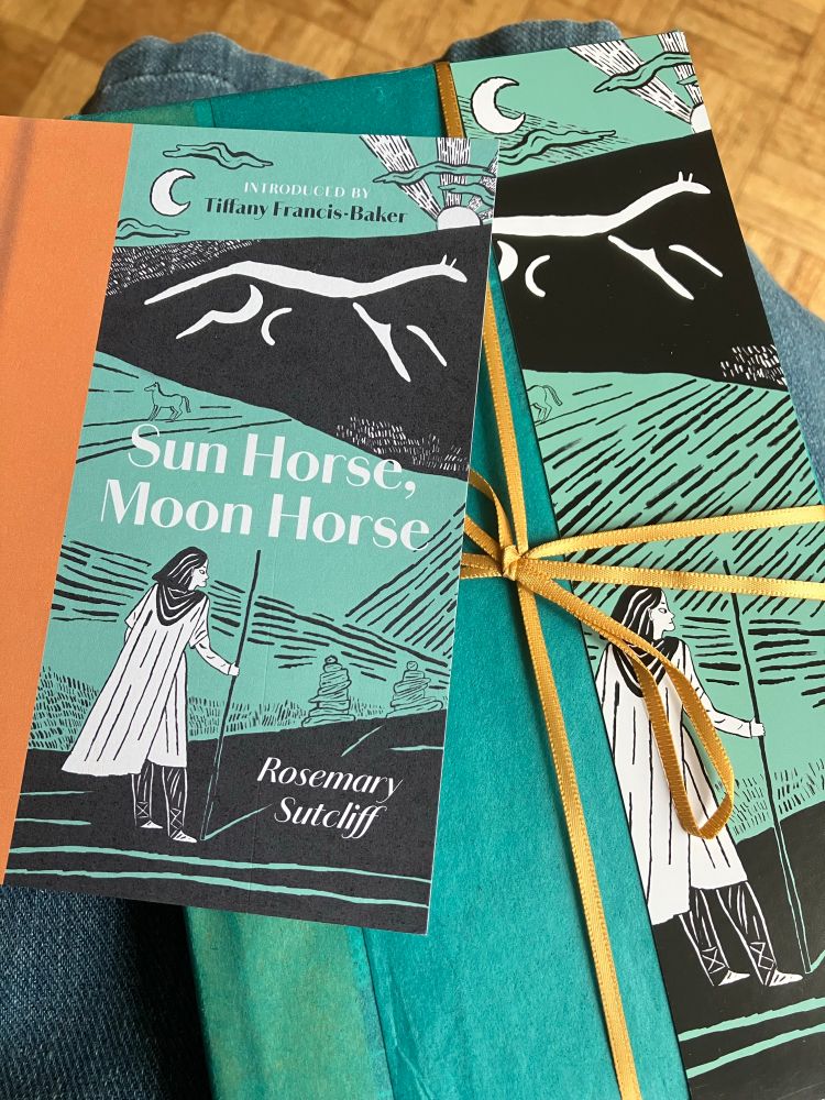 The Manderley Press edition of Rosemary Sutcliff’s ‘Sun Horse, Moon Horse’ (introduced by Tiffany Frances Baker) — perfectly gift-wrapped in turquoise tissue paper and tied with a saffron ribbon. There’s a matching bookmark and postcard with the book’s cover design (by Isabel Greenberg) of a man in tunic, leggings and cloak looking up at the White Horse of Uffington, beneath the sun and a crescent moon.