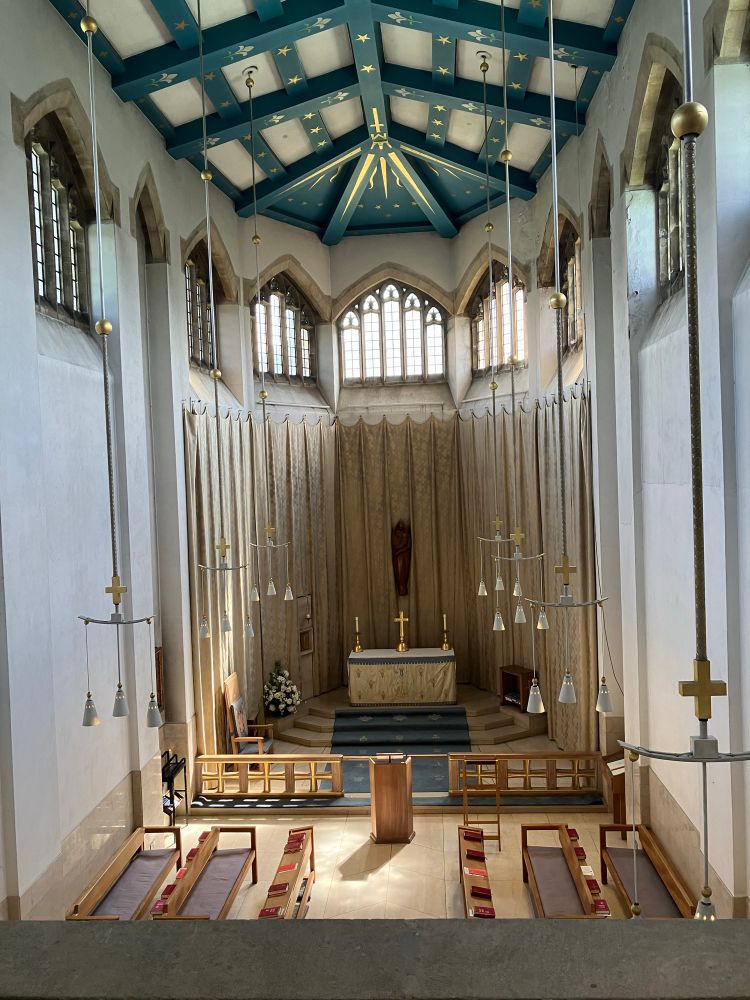 View of the Lady Chapel from high up at the west end. The building is of pale cream stone and the windows have clear glass. The ceiling is also cream but with bands painted teal with gold stars; minimalist white and gold lamps hang from it on long, pale cords. There is a modern wooden carving of the Virgin and Child above the altar, which bears a gold-and-red cross and matching candlesticks and has a pale gold frontal. The sanctuary carpet is pale grey-blue with faded gold decorations. Seating consists of pale wooden benches with grey upholstery. 