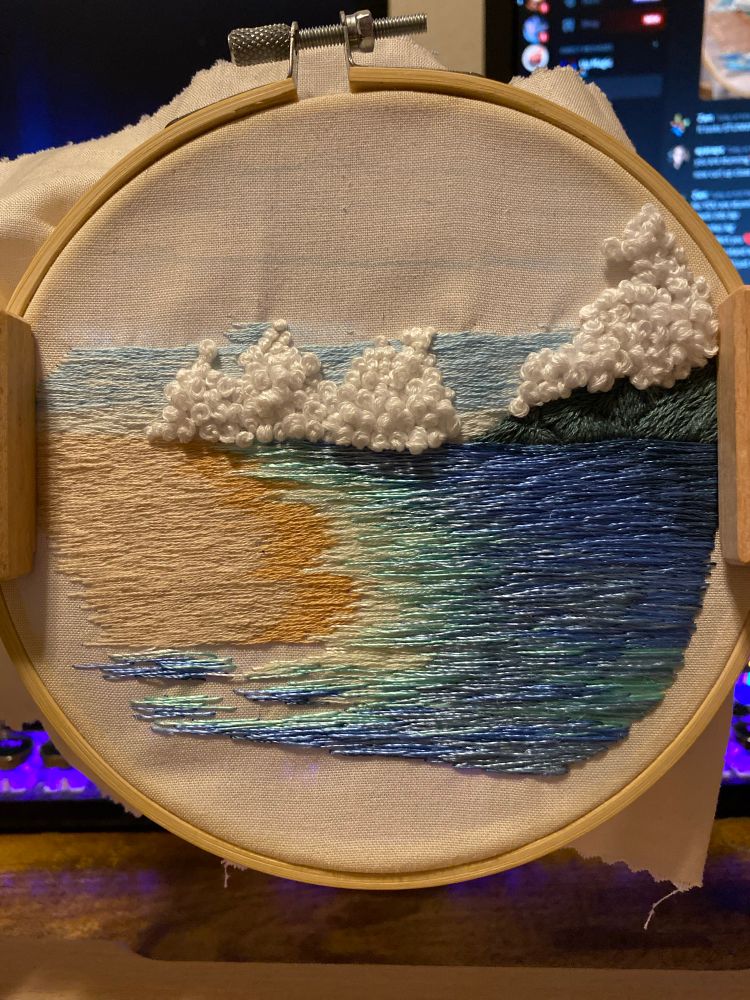 partially embroidered scene of a beach with waves coming in, clouds roll in from the distance, and behind them slowly a blue sky is being stitched in. Very slowly. 