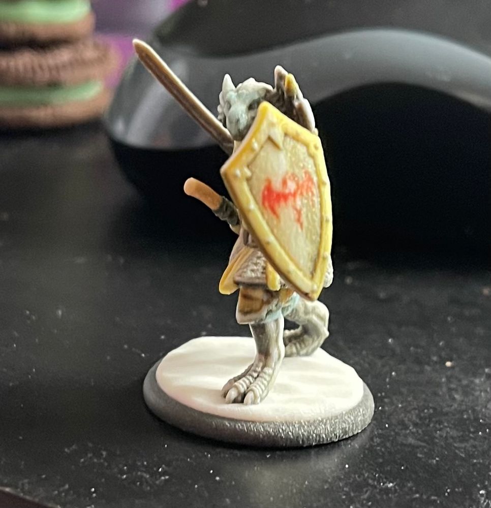 A sliver Dragonborn mini figure standing on a snowdrift base, clad in chain mail with yellow-ish accents, wielding a long sword and shield. A hand axe is on his belt, and a battle axe is strapped to his back. He is posed in a defensive battle stance. His shield has a draconic emblem painted on it. 