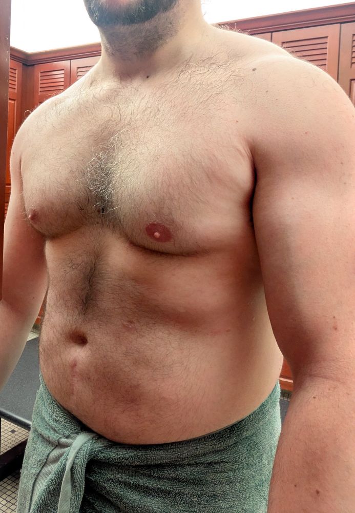 A picture of me shirtless with a towel wrapped around my waist, turned towards the camera to show off my torso. The photo is cropped so that my face is not visible besides my short beard, and my legs and below are out of frame as well. I am illuminated by a strong light from directly above, which I feel is particularly flattering.