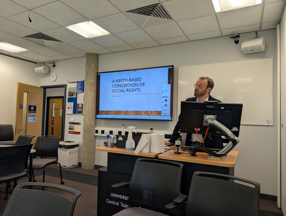 Picture of Marco Goldoni behind a lecture lectern in a university room, with power point presentation with the title of the talk in the background.