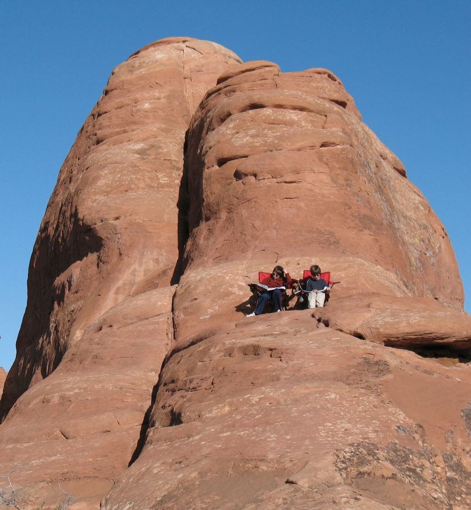 two children in red folding chairs, sitting halfway up a red sandstone cliff, reading Calvin and Hobbes 
