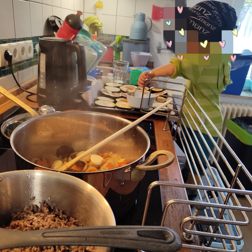 Blick über den Herd in die Küchenzeile. Auf dem Herd Töpfe. Im Hintergrund ein Kind mit verpixeltem Gesicht, das Auberginenscheiben bestreicht.