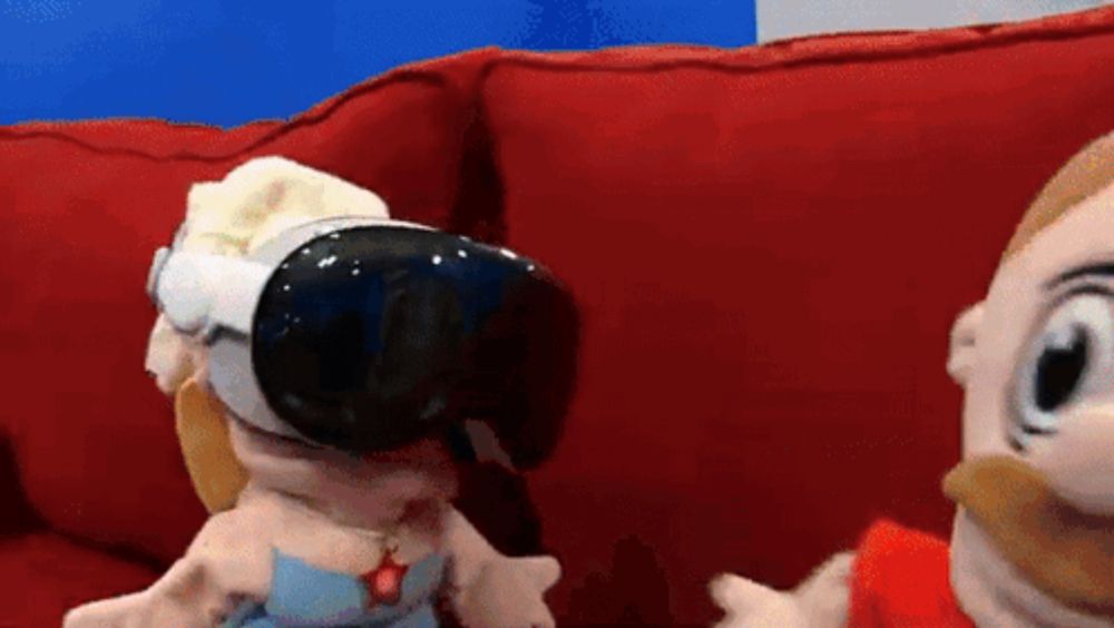 two stuffed animals are sitting on a red couch and one is wearing a virtual reality headset