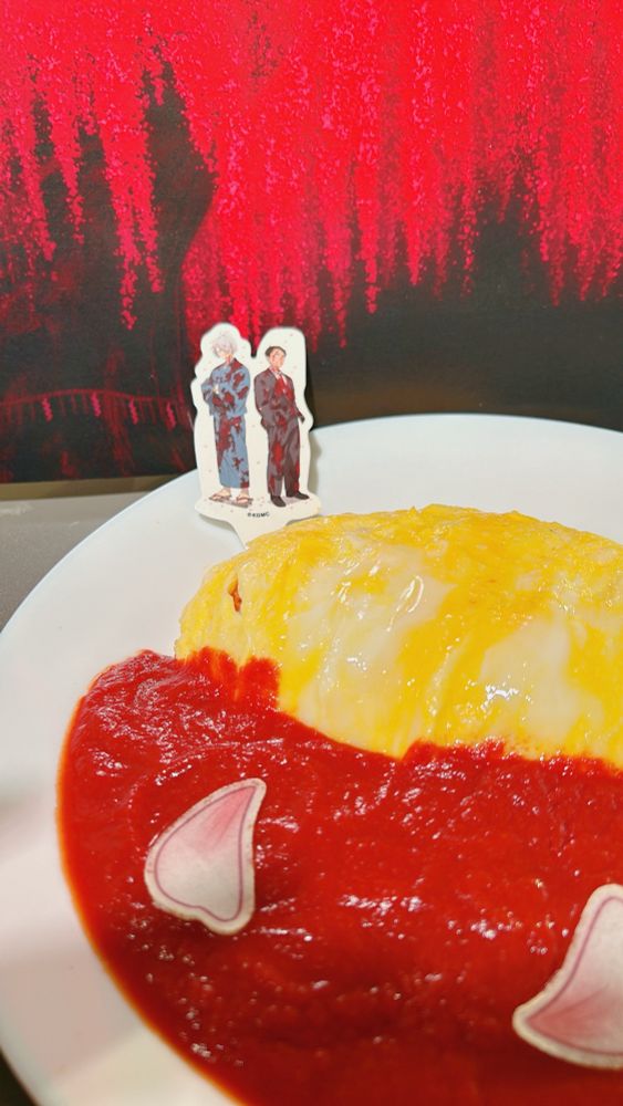 ゲ謎のコラボカフェの血桜オムライスの写真。大量の赤いソースが血の池のよう。血まみれでかっこよく佇む父たちが背中合わせになっている絵が描かれた飾りがオムライスの端に刺さっており、皿の向こうにはおどろおどろしい血桜の物撮り用背景が写っている。