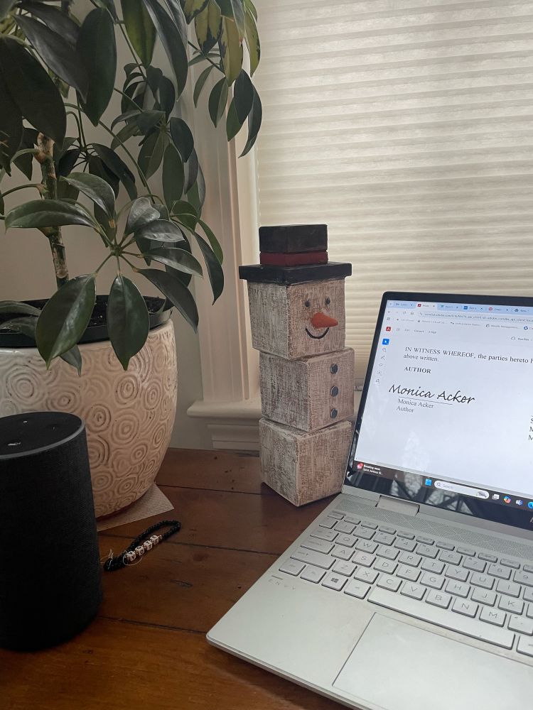On a wooden table sits a silver computer opened to a screen that shows the signing of a contract with the words “Monica Acker - Author.” Beside the computer is a happy snowman made of white-painted wooden blocks, a tall green plant in a white, textured planter, a child’s beaded bracelet, and an Alex device. 