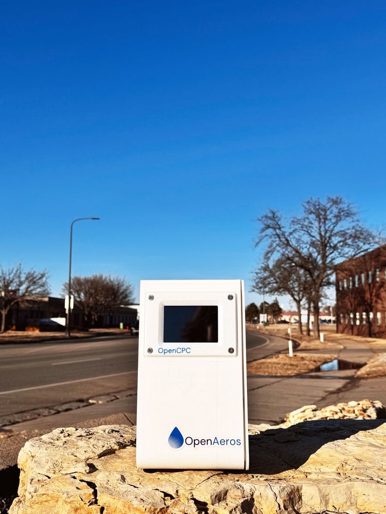 A small, white prototype OpenCPC(TM) with the OpenAeros(TM) logo sits on a flagstone outside against, well, a big ol' blue sky. 
