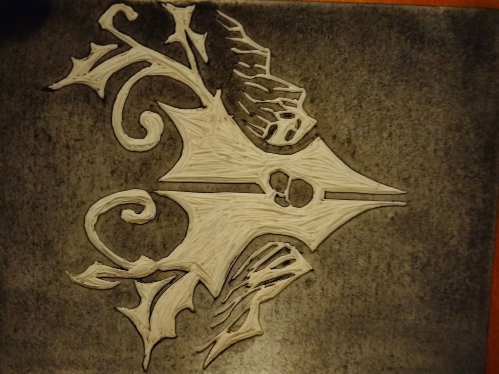 A carved rubber block with ink on it. The design is the Mount Holly logo from Blue Prince, of a piece of holly with mountains behind it