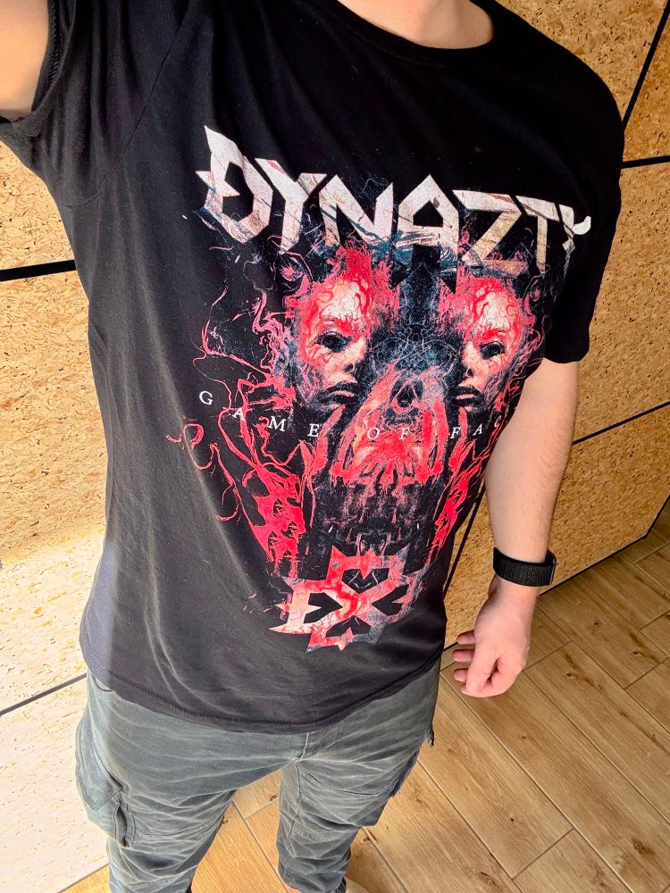 Selfie vom Outfit vom Hals bis etwas unter die Knie. Ich trage neben einer schwarzen Cargo Hose und einer Smartwatch mit dunkelgrauem Uhrband ein Bandshirt von Dynazty auf dem das Bandlogo als Wortmarke oben steht ganz unten eine rot schattiertes Logo ist, das an ein eckiges vierblättriges Kleeblatt erinnert in der Mitte sind zwei halbe Gesichter in rot-orangenen Tönen schattiert und ein nicht näher definierterbares Muster mit auslaufenden in geschwungenen Linien. Darüber steht Game of faces in Großbuchstaben mit größerem Abstand zwischen den Zeichen
