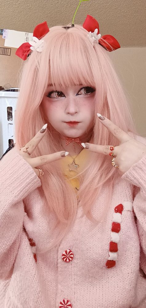 Young person with pink hair, candy-themed sweater, and red bows makes playful peace signs. They exude a whimsical, cute aesthetic.