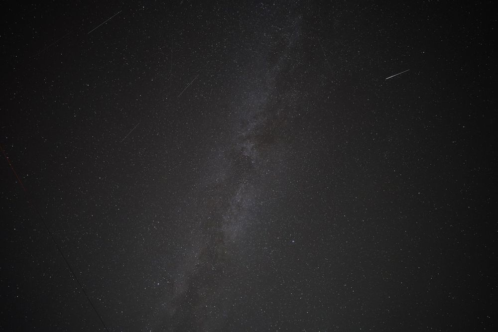 Looking straight up at the Milky Way, stretching diagonally across the photo, with a few satellite streaks at random angles throughout the whole photo, including one that's glinting very brightly. 