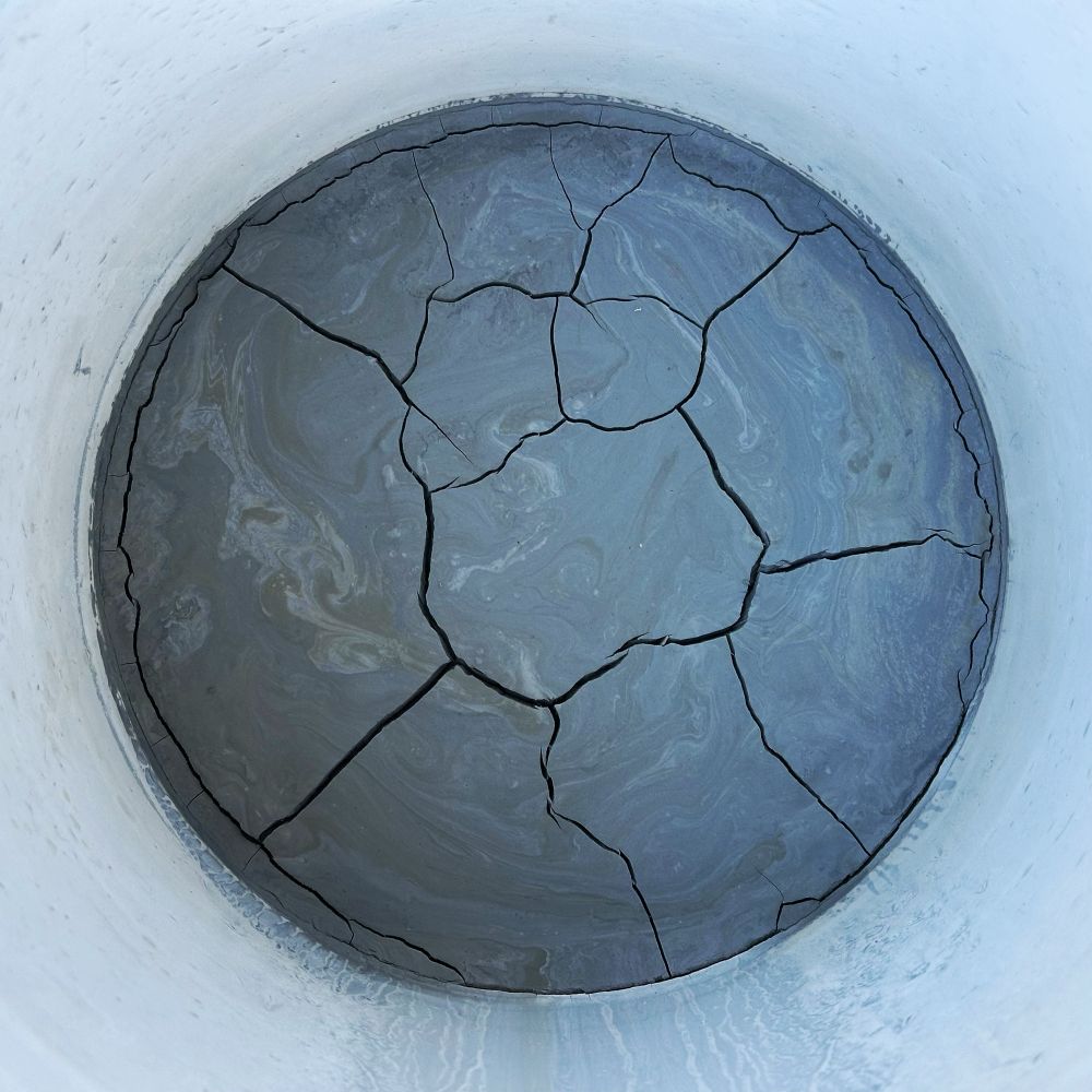 A white bucket containing dried rock tumbler slurry, a combination of grit and rock dust. The mass has swirls and desiccation cracks. This is good (I left it out in the Sun) as I can just scrap it into the trash.
