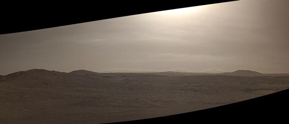 A desolate terrain with craters and distant buttes. Thin clouds are visible in the sky.