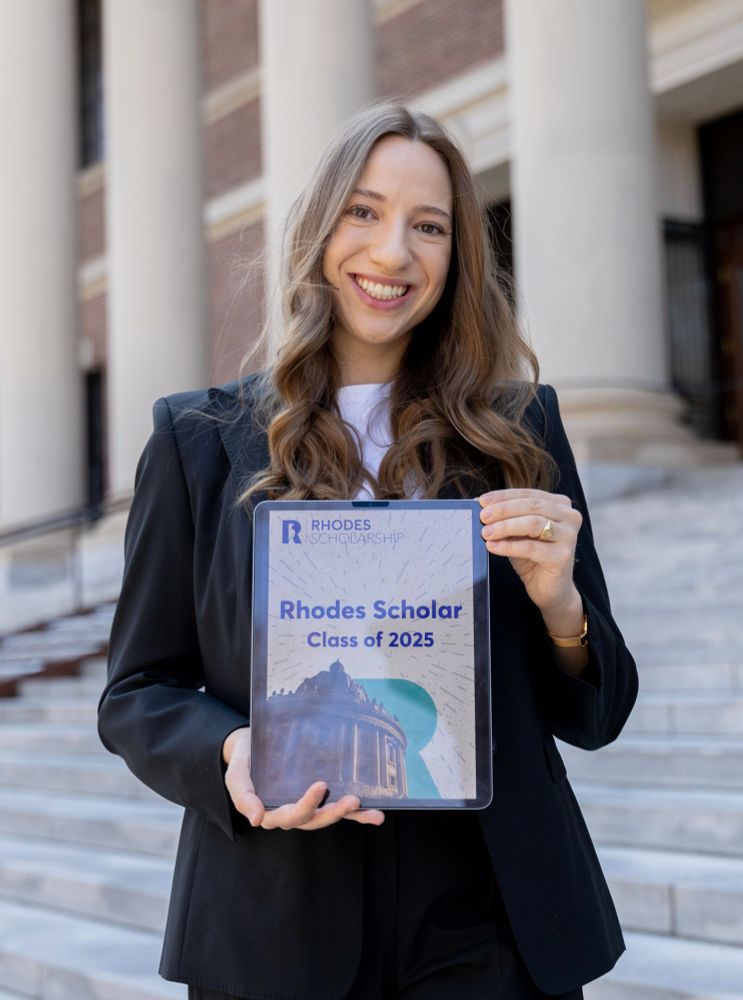 Laura standing on steps holding up the Rhodes scholar selection poster on an iPad