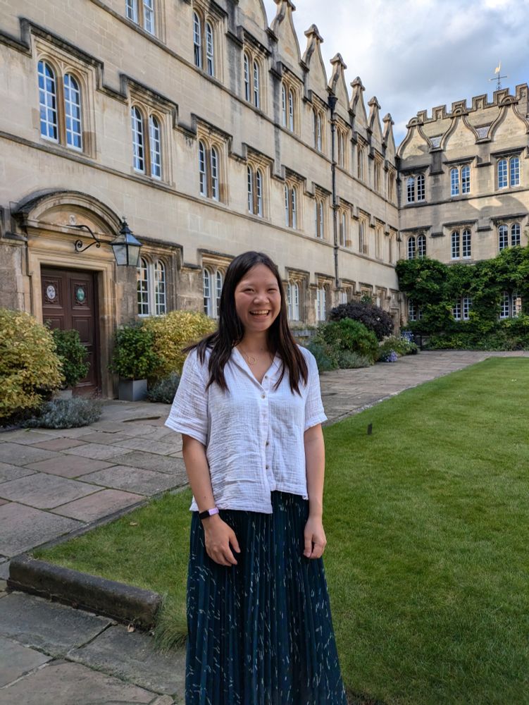 Zandy wearing a white shirt and navy skirt, standing in a quad in Jesus College next to grass and old buildings
