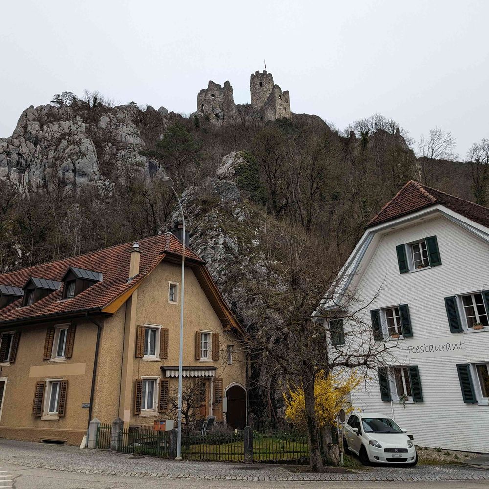 Bild einer Burgruine hoch auf einem Felsen. Im Vordergrund zwei Häuser unten im Tal.