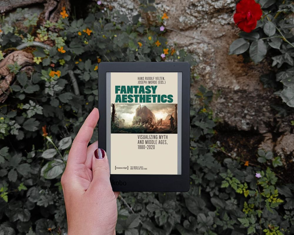 a white hand holds an e-reader showing the cover of the book in front of a stone wall covered in greenery.