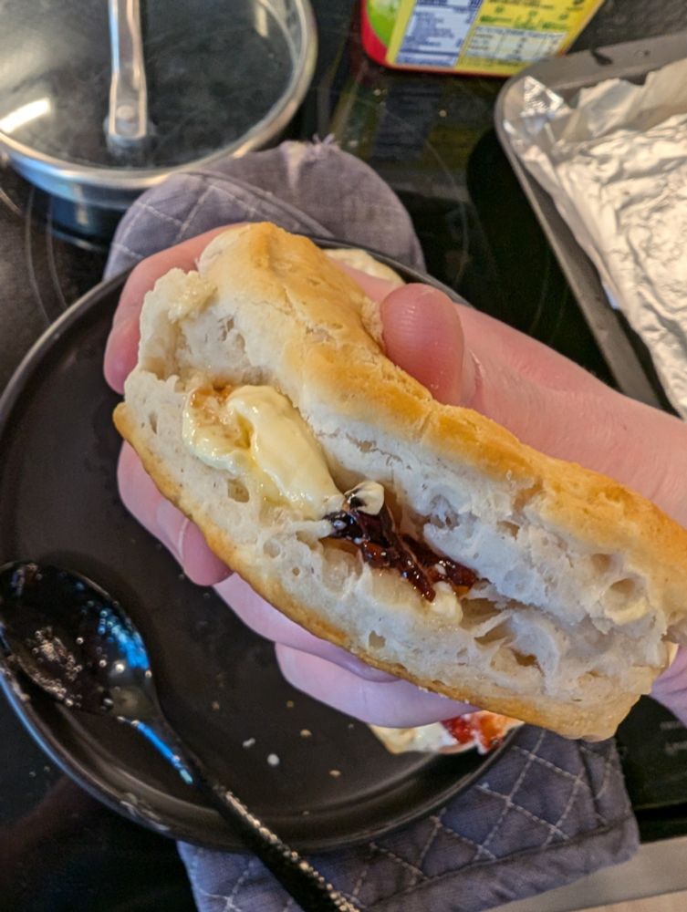 one biscuit sandwich filled with butter and jam, the previous photos plate is the backdrop with other cookinf implements spread about
