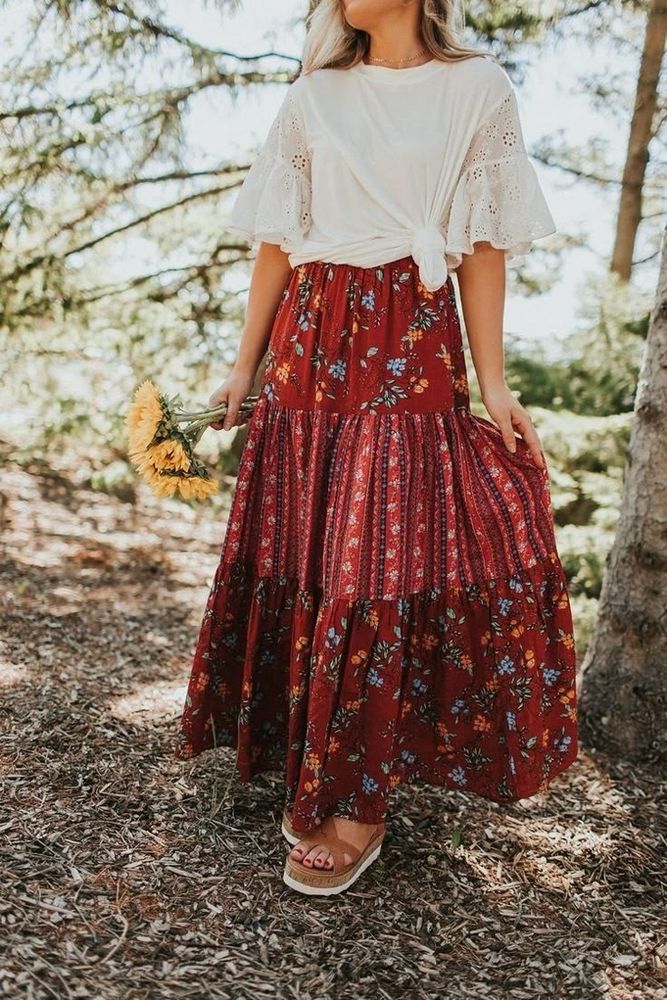 A woman wearing a modest white top with lacy flared 3/4 sleeves, tied into a crop top style, with a red patchwork flowy maxi skirt and platform sandals. 