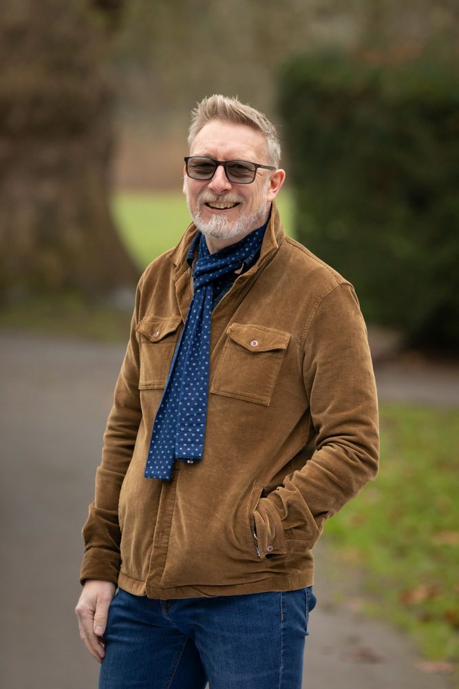 A man with short blonde/grey hair and a beard wearing spectacles with blue lenses, wearing a brown jacket, jeans and a blue scarf stands in a rural backdrop