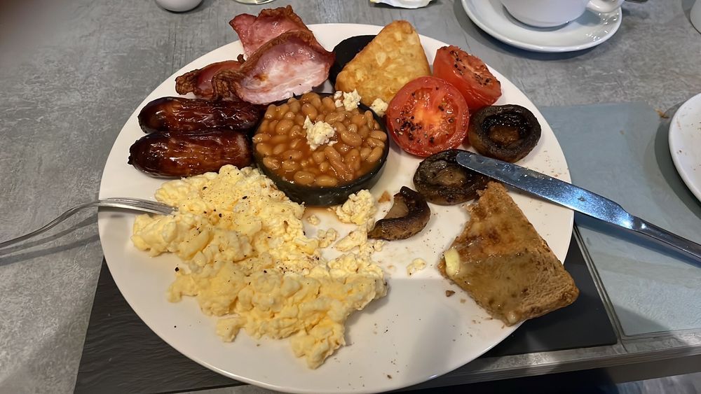 A picture of a breakfast plate full of bacon rashers, sausages, black pudding, scrambled eggs, mushrooms, hash brown, beans, toast and grilled tomatoes 