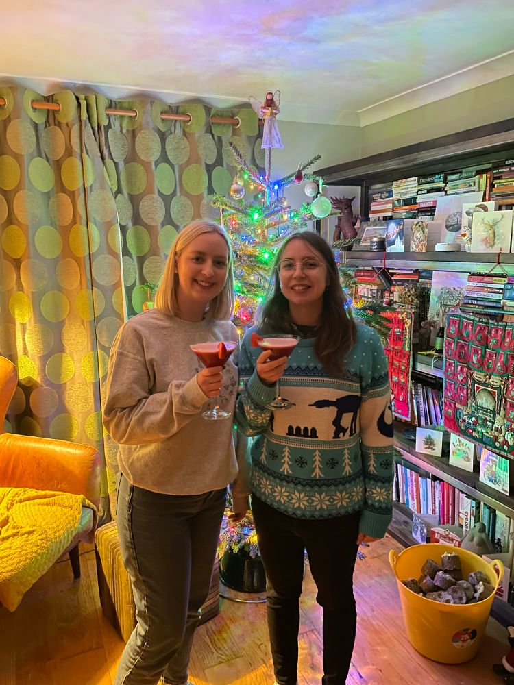 Two women with cocktails in hand, one with a Christmas jumper on, stand in front of a Christmas tree all lit up.