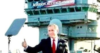 The image is a photo of George W. Bush giving a thumbs up during his famous Mission Accomplished speech from the deck of USS Abraham Lincoln on May 1st, 2003, with the Mission Accomplished banned behind him. George W. Bush served as the 43rd US President. He currently resides in McLennon County, Texas, USA