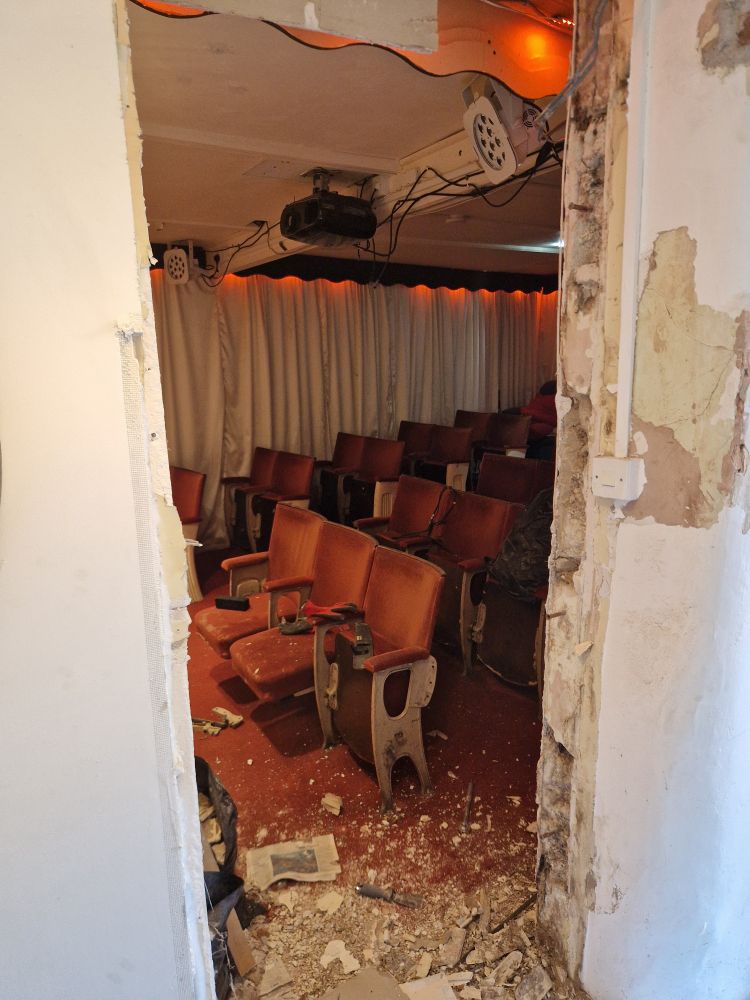 A hole in a wall, revealing some vintage cinema seats. The masonry/rock wall that stands to the right was the source of water ingress - and cement render gave the water nowhere else to go except into the stud wall, where plasterboard, gypsum plaster and PIR board kept the water within. The rot was inevitable. 