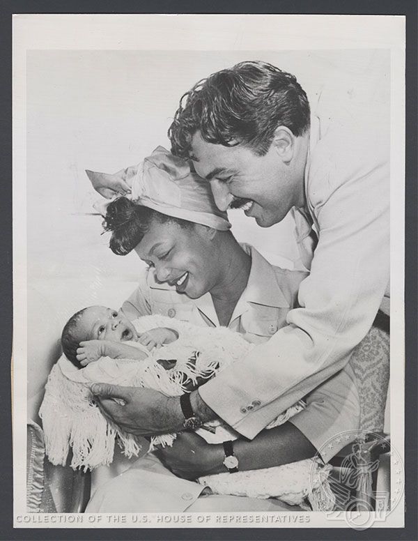Adam Clayton Powell Jr. smiles and leans over Hazel Scott, seated, who smiles and holds their newborn son