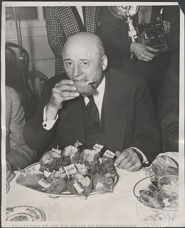 Speaker Rayburn eats a potato in front of a large plate of potatoes