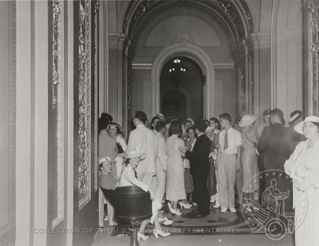 1930s photograph of Capitol visitors crowded in a hallway. Many are wearing white shoes and straw hats.