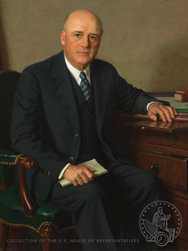 A balding man sits in a chair in a dark suit and blue stripe tie. He has a serious expression on his face.