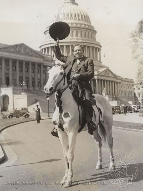 A man in a suit on a white horse on the East front of the Capitol waves his hat.