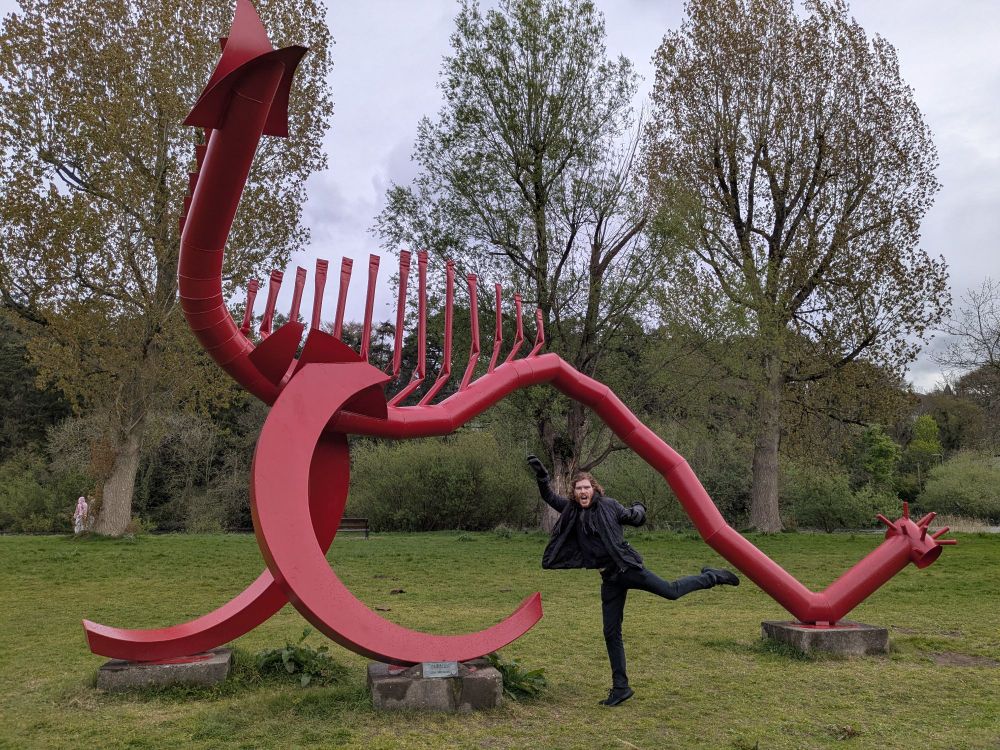 A man standing in front of a large, red, metal sculpture which kind of looks like a stegosaurus (the sculpture not the man) with trees in the background