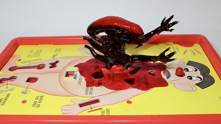 A toy Xenomorph bursting out of an Operation board game, with red goo spread across simulating blood.