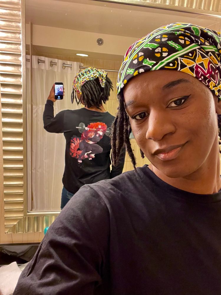 Maya posing with her face showing and her back angled to the mirror. The back of the shirt has a picture of an anthropomorphic tiger woman who is weightlifting mid squat with long red hair and black fitness clothes. Maya is wearing makeup and a multicolored bandanna on here head. 