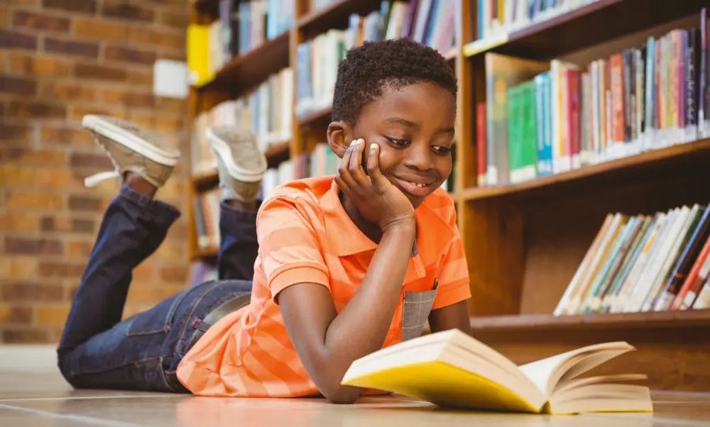 A boy lying on the floor reading a book with a bookshelf beside him. 