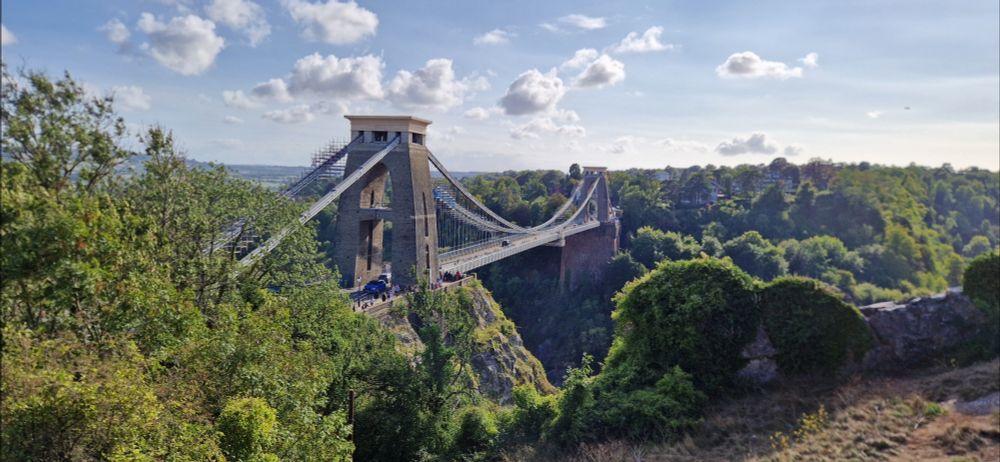 View of Clifton Suspension Bridge from the Clifton Observatory. The bridge is framed by luscious green trees and there is a beautiful blue sky, with few clouds