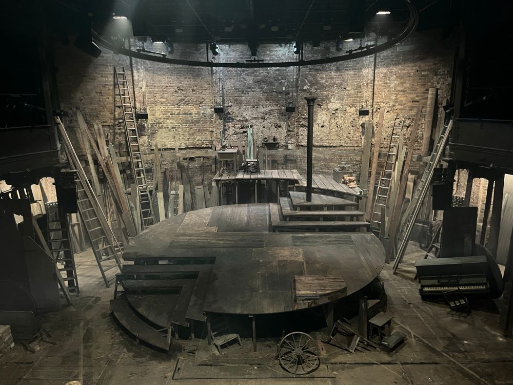 View of the stage for A Moon for the Misbegotten at Almeida Theatre featuring a raised round stage made of wood surrounded by a clutter of planks, ladders and derelict items. 