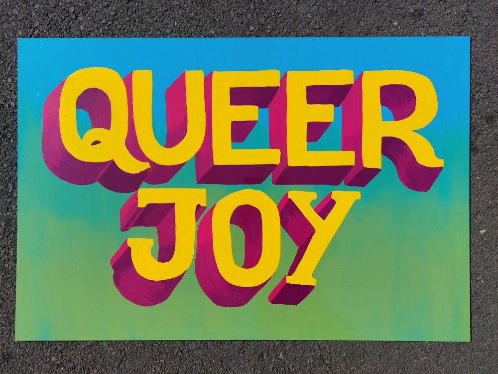 A painted dibond sign on a wet tarmac surface. The sign says "QUEER JOY" in yellow capital letters with purple blocks, and the background is a pale blue to green gradient.