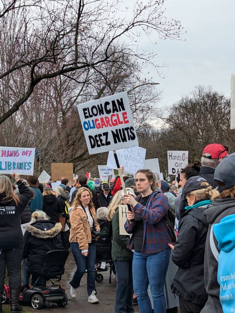 Posters at Rally. Sign reads Elon Van OLIGARGLE Deez Nuts.