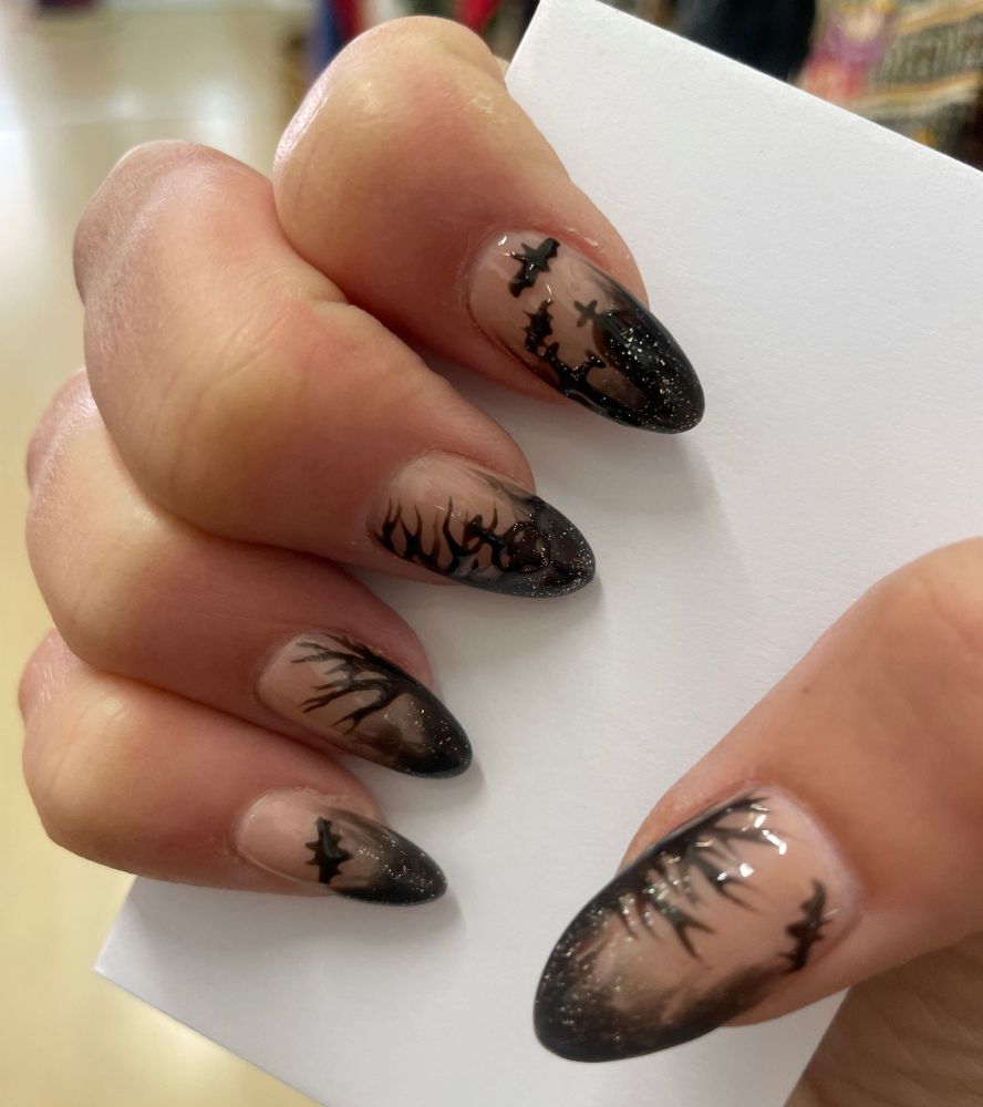 right hand clasped around a white envelope showing my manicure, halloween/spooky season inspired, black french tip blend back, looking shadowy, into a natural base colour, each nail has a different hand painted design on it, bats, trees, headstone from a grave, & a high cross, then finishes with silver glitter over the black tip. 