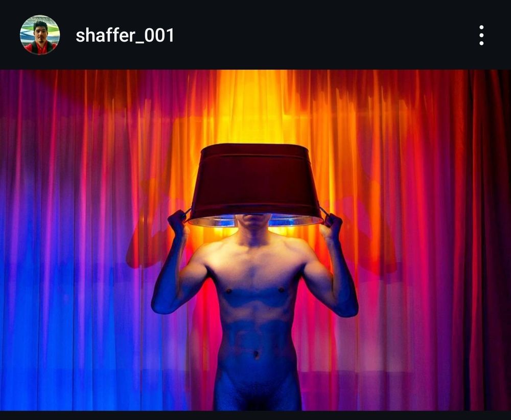 Homem nu, de frente pra câmera, segurando o que parece ser uma champanheira grande enfiada na cabeça, como se fosse a parte de cima de um abajur vermelho escuro. Atrás dele há uma cortina iluminada em tons que vão do roxo e azul até o vermelho e vinho, passando por amarelo e laranja.