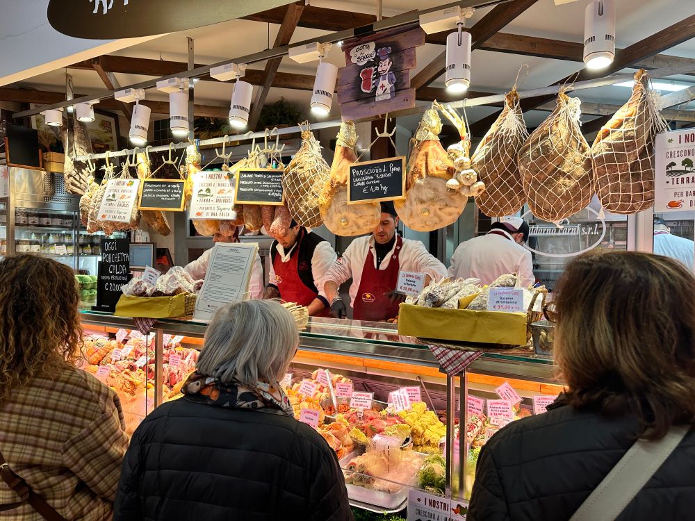 A meat stall with prosciuttos hanging from the ceiling and many local Florentine ladies lining up 